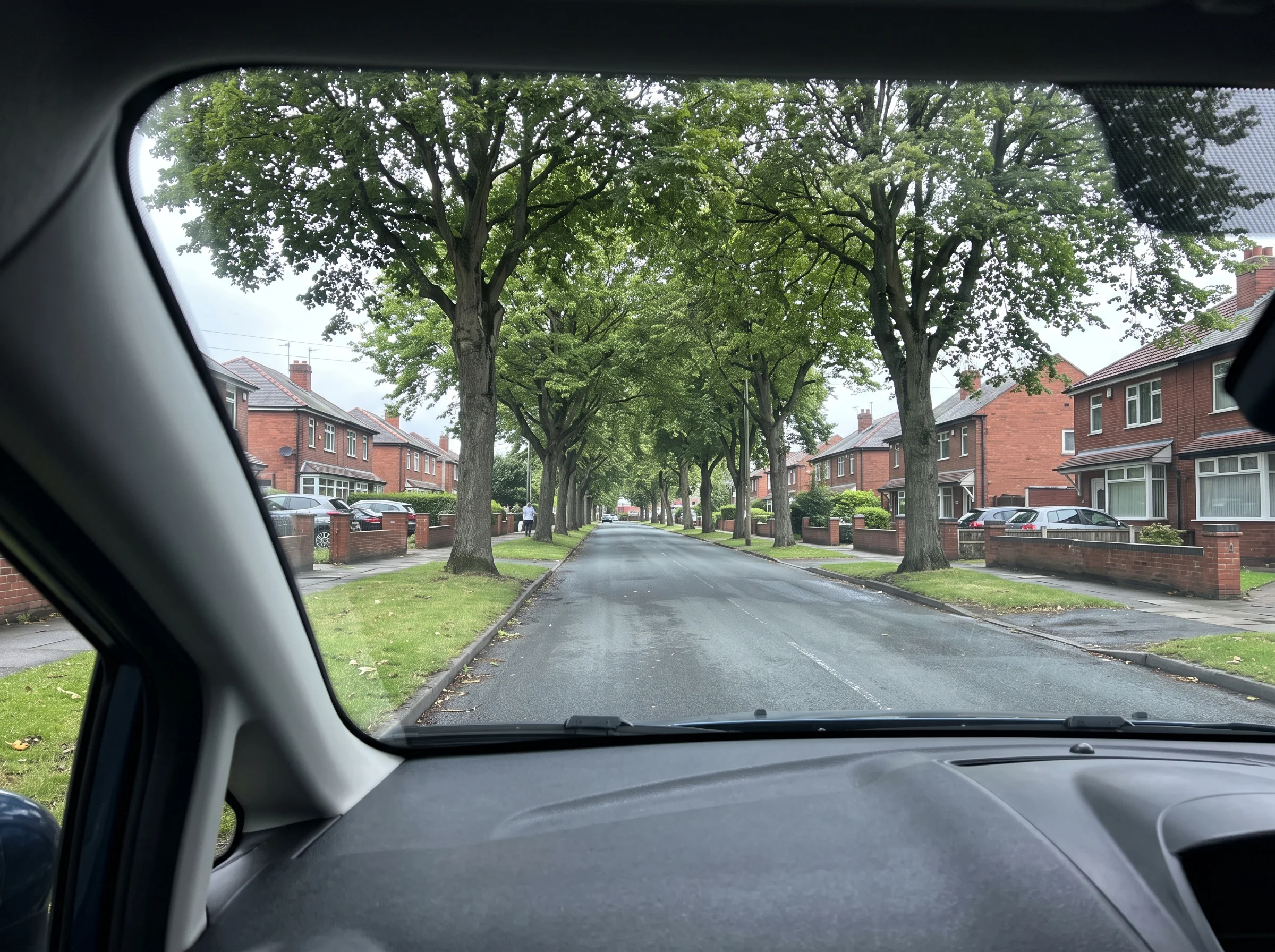 A quiet Birmingham residential road lined with trees and red-brick houses, seen through a car windscreen on an overcast morning